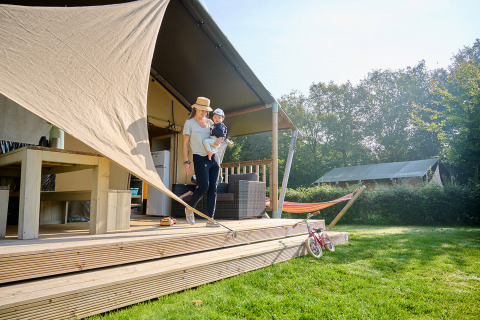 Madre con niño en la terraza frente a una tienda en Camping de Papillon, Overijssel, Países Bajos, día soleado.