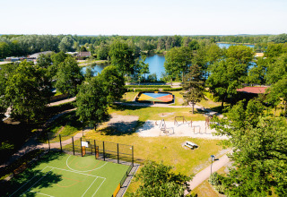 Aerial view of Camping de Schatberg with a playground, sports court, and lake surrounded by trees in Limburg.