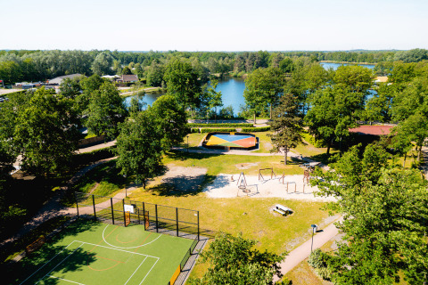 Aerial view of Camping de Schatberg with a playground, sports court, and lake surrounded by trees in Limburg.