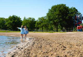 Twee meisjes wandelen langs het meer op Camping de Schatberg, Limburg, Nederland, met glijbaan in zicht.