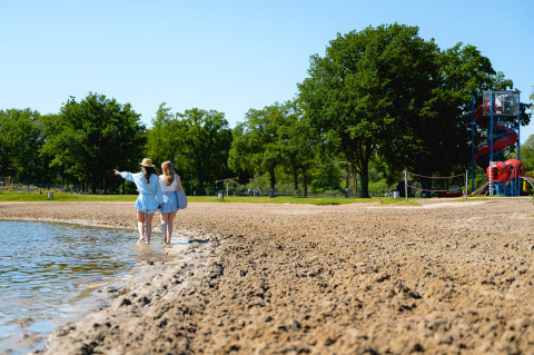 Two girls walk along the lakeshore at Camping de Schatberg in Limburg, Netherlands, with slide in the distance.