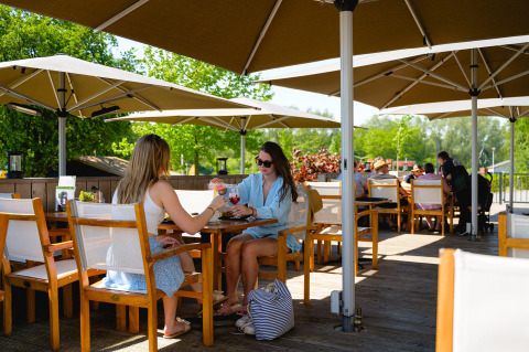 Two women enjoy drinks on a sunny terrace under large umbrellas at Camping de Schatberg, Limburg.