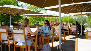 Dos mujeres disfrutan de bebidas en una terraza soleada bajo sombrillas en Camping de Schatberg, Limburg.