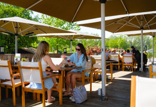 Dos mujeres disfrutan de bebidas en una terraza soleada bajo sombrillas en Camping de Schatberg, Limburg.