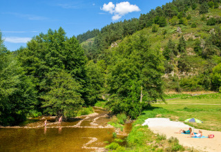 Gezinnen ontspannen aan een rivierstrandje bij Camping de Vaubarlet, omgeven door groene heuvels in Frankrijk.
