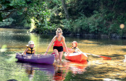 An adult helps two children in kayaks on a shallow river at Camping de Vaubarlet, Auvergne-Rhône-Alpes, France.
