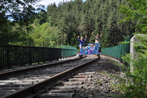 Famiglia in draisina attraversa un ponte ferroviario nel verde al Camping de Vaubarlet, Auvergne-Rhône-Alpes, Francia.