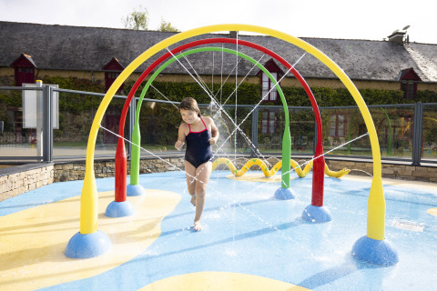 Niño jugando bajo arcos de agua de colores en el parque infantil de Camping Domaine du Logis, Bretaña, Francia.