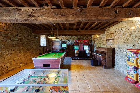 Sala de juegos con paredes de piedra, billar, futbolín, máquinas recreativas y piano en el camping.