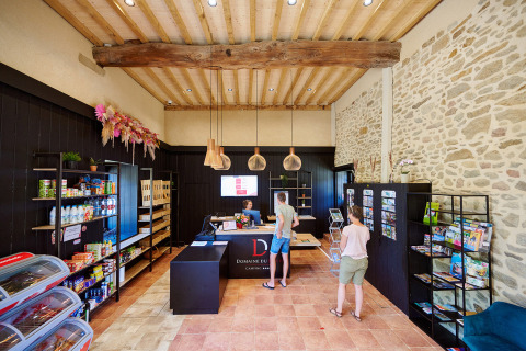 Interior de la recepción y tienda en Camping Domaine du Logis, con visitantes y expositores de folletos informativos.