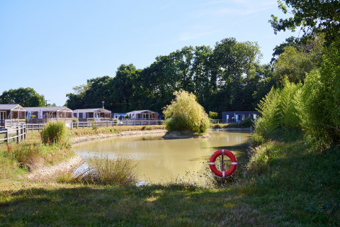 Parque vacacional con cabañas junto a un estanque en Camping Domaine du Logis, Bretaña, Francia, rodeado de árboles.