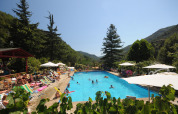 Udendørs swimmingpool omgivet af solsenge og parasoller i Camping Delle Rose, Ligurien, Italien, med bjerge.