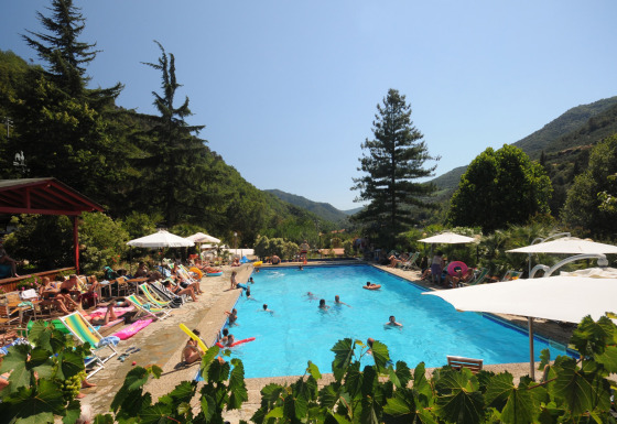 Udendørs swimmingpool omgivet af solsenge og parasoller i Camping Delle Rose, Ligurien, Italien, med bjerge.