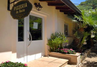 Ingang van Pizzeria da Bairò met bloemen en palmbomen, genomen bij Camping Delle Rose in Ligurië, Italië.