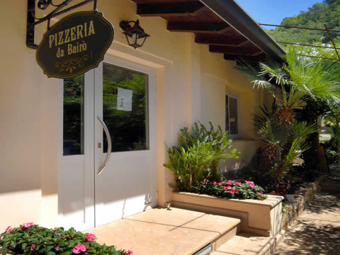 Entrée de la Pizzeria da Bairò avec fleurs et palmiers au Camping Delle Rose, parc de vacances en Ligurie, Italie.