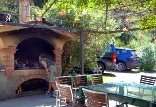 A man checks food cooking in an outdoor stone oven at Camping Delle Rose, with tables and parked cars nearby.