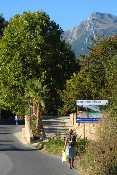 Kvinde går på vej mod Camping Delle Rose i Ligurien, Italien, med træer og bjerge i baggrunden.
