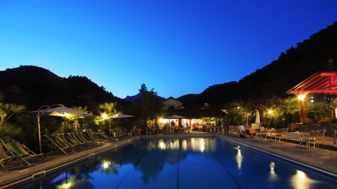 Piscina iluminada en Camping Delle Rose, un parque vacacional en Liguria, Italia, al atardecer entre montañas.