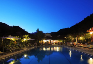 Piscina illuminata a Camping Delle Rose in Liguria, Italia, circondata dalle montagne in un'atmosfera serale.