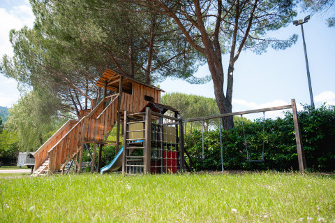 Parque infantil con casa de madera, tobogán y columpios bajo altos pinos en Camping Il Poggetto, Toscana, Italia.