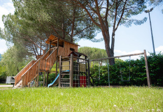 Speelplein met houten huisje, glijbaan en schommels onder dennen bij Camping Il Poggetto in Toscane, Italië.