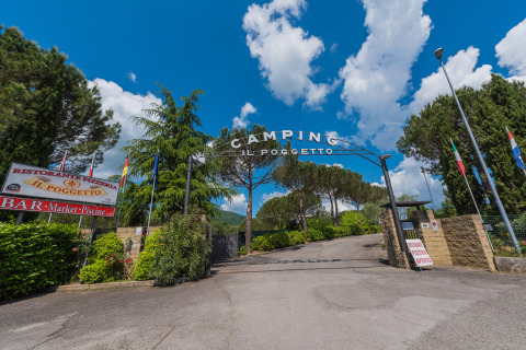 Ingresso del Camping Il Poggetto, villaggio vacanze in Toscana, Italia, con alberi e cielo azzurro.