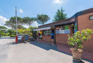 Réception du Camping Il Poggetto en Toscane, Italie, avec un visiteur, des panneaux et végétation verdoyante.