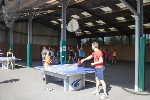 Bambini e adulti giocano a ping pong e basket in un’area sportiva coperta al Camping La Bretonnière.