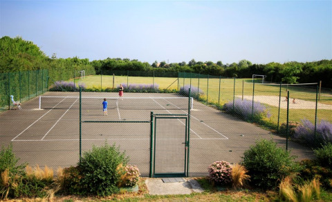 Two people playing tennis on a fenced court with flowers and a grassy field at Camping La Bretonnière, France.