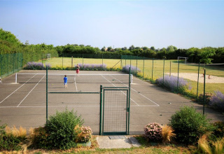 Two people playing tennis on a fenced court with flowers and a grassy field at Camping La Bretonnière, France.