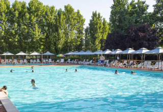 Udendørs swimmingpool med folk der svømmer og mange liggestole under parasoller, omgivet af grønne træer.