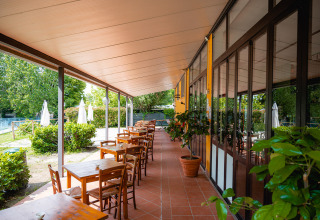 Terrazza coperta con tavoli e sedie in legno, piante in vaso e vista giardino al Camping La Chiocciola in Toscana.