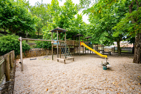 Parque infantil con columpios, tobogán y trepador en Camping La Clémentine, Occitania, Francia, rodeado de árboles.