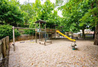 Playground with swings, a slide, and climbing frame surrounded by trees at Camping La Clémentine, Occitanie, France.
