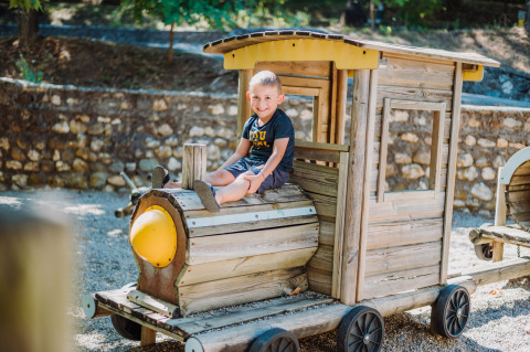 Un bambino sorride seduto su un trenino di legno nell'area giochi di Camping La Clémentine, Occitania, Francia.