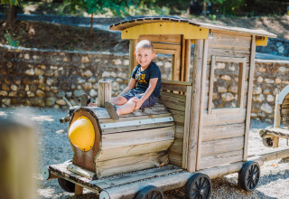 Un bambino sorride seduto su un trenino di legno nell'area giochi di Camping La Clémentine, Occitania, Francia.