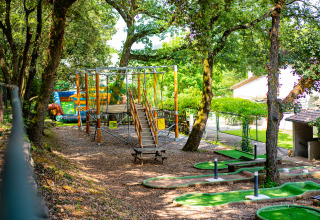 Área de juegos y minigolf entre árboles en Camping La Clémentine, parque vacacional en Occitanie, Francia.