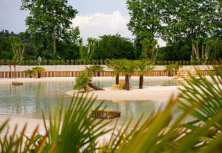 Camping La Clémentine holiday park in Occitanie, France, with palms, sandy shore, and tranquil water.