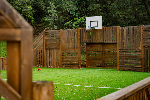 Campo sportivo con canestro da basket e prato sintetico al Camping La Clémentine, Occitania, Francia, recinto in legno.