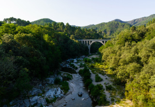View of lush green forest, river, and stone arch bridge at Camping La Clémentine in Occitanie, France.
