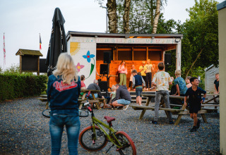 Børn og voksne samles ved udendørs scene i Camping La Colline, Belgien, med legecykel i forgrunden.