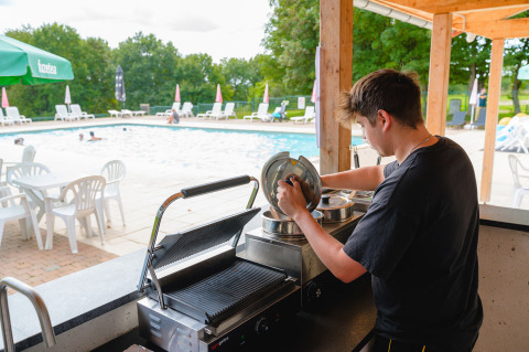 Una persona prepara comida en un bar junto a la piscina en Camping La Colline, parque de vacaciones en Bélgica.