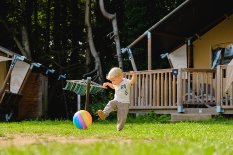A young child kicks a colorful ball on the grass in front of a wooden cabin at Camping La Colline in Belgium.