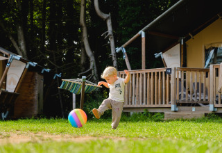 A young child kicks a colorful ball on the grass in front of a wooden cabin at Camping La Colline in Belgium.
