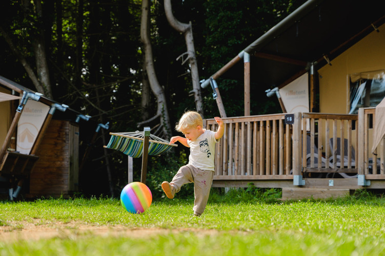 Ein kleines Kind kickt einen bunten Ball auf der Wiese vor einer Holzhütte im Campingpark La Colline, Belgien.