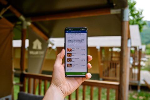 Persona sosteniendo un móvil con una app de pedidos frente a cabañas en Camping La Garenne, Francia.