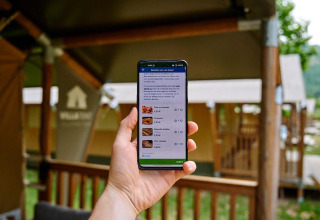 Eine Person hält ein Smartphone mit Bestell-App vor Ferienhütten im Camping La Garenne, Frankreich.