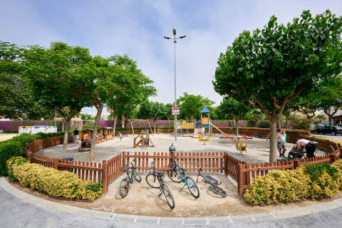 Parque infantil vallado en Camping La Masia, Cataluña, España, con bicicletas, árboles y niños jugando.