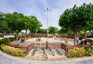 Afgebakende speeltuin met fietsen, bomen en spelende kinderen bij Camping La Masia, Catalonië, Spanje.