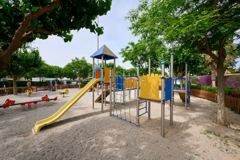 Parque infantil con tobogán y estructura para escalar en Camping La Masia, rodeado de árboles y valla.